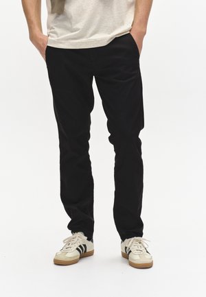 Schwarze Hose im Slim-Fit, aus Baumwollstoff, mit seitlichen Taschen. Kombiniert mit cremefarbenen Sneakers mit schwarzen Streifen.