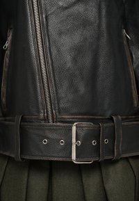 Černá kožená bunda s texturovaným povrchem, která má stříbrný zip, široký pásek s kovovou přezkou a tmavé švy.