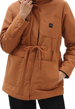 Personne portant une veste marron avec des poches avant, une taille à cordon de serrage et un patch avec le logo Vans sur la poche poitrine.