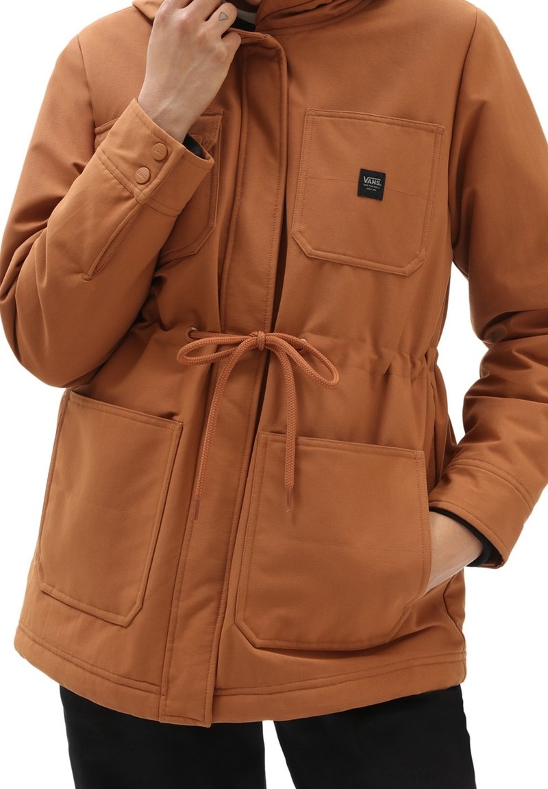Personne portant une veste marron avec des poches avant, une taille à cordon de serrage et un patch avec le logo Vans sur la poche poitrine.