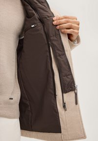 Beige wollen jas met een bruin satijnen interieur, voorzien van een ritszak en zilveren hardware. De jas heeft een gladde textuur en een getailleerd ontwerp.