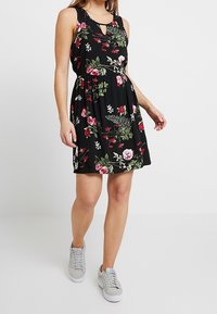 Svart ärmlös klänning med blommönster i rosa och gröna accenter, midja som är cinchad och en nyckelhålsdetalj vid halsringningen.