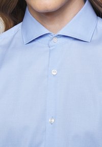 Gros plan d'une personne portant une chemise bleu clair texturée à boutons avec un col pointu, boutons fermés en haut et au milieu.