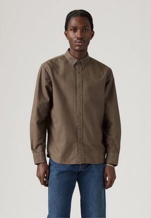 Junger Mann mit geflochtenem Haar, der ein braunes Hemd mit Knöpfen und eine blaue Jeans trägt, vor einem einfachen hellen Hintergrund stehend.