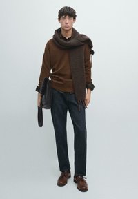 Brauner Pullover mit einem dicken, dunklen Schal, der über die Schultern gelegt ist, dunkle Jeans und braune Lederschuhe. Der Schal ist mit Fransen versehen.