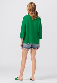 Vrouw met golvend haar, gekleed in een groene lange mouwen top, versleten denim shorts en groene open achterkant sandalen, staand met haar rug naar de camera gekeerd.