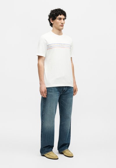 Νέος άνδρας με σγουρά μαλλιά, φορώντας λευκό μπλουζάκι με λεπτές οριζόντιες ρίγες, φαρδιά μπλε τζιν και ανοιχτό καφέ μοκασίνια.