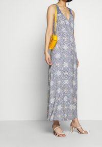 Robe maxi fleurie bleu avec un décolleté plongeant, présentant un design à motifs. Accompagnée d'un sac à main jaune et de sandales à talons nudes.