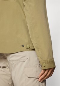 Veste olive claire au toucher lisse, avec des pressions en bas. Associée à un pantalon cargo beige avec des poches à rabat.