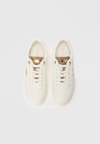 Parillinen valkoisia matala-top sneaker-kenkiä, joissa on ruskea käärmeennahkatekstuuri kielessä ja kultaiset logo-yksityiskohdat valkoisella taustalla.