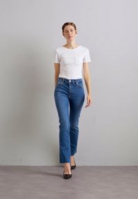 White short-sleeve t-shirt with pocket, blue high-waisted jeans, and black pointed-toe heels. Model walks against a neutral background.