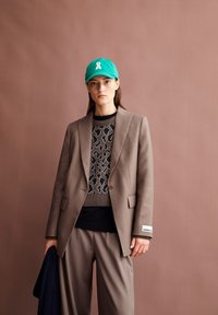 Vrouw in een taupekleurig pak met een trui met luipaardprint en een groene pet, die een marineblauw kledingstuk vasthoudt tegen een effen, gedempt bruine achtergrond.