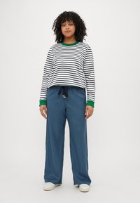Camicia a righe con maniche lunghe e polsini verdi, abbinata a pantaloni blu con coulisse larghi e scarpe chiare.