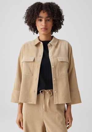 Frau mit lockigem Haar trägt eine beige Cordjacke mit Knöpfen, passende Hose mit Tunnelzug und darunter ein schwarzes, geripptes Top.
