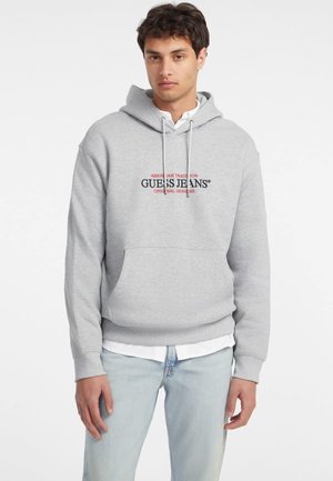 Grijze hoodie met trekkoord, voorzien van een zwart en rood logo op de borst. Draag het over een witte blouse en combineer het met lichtblauwe jeans.