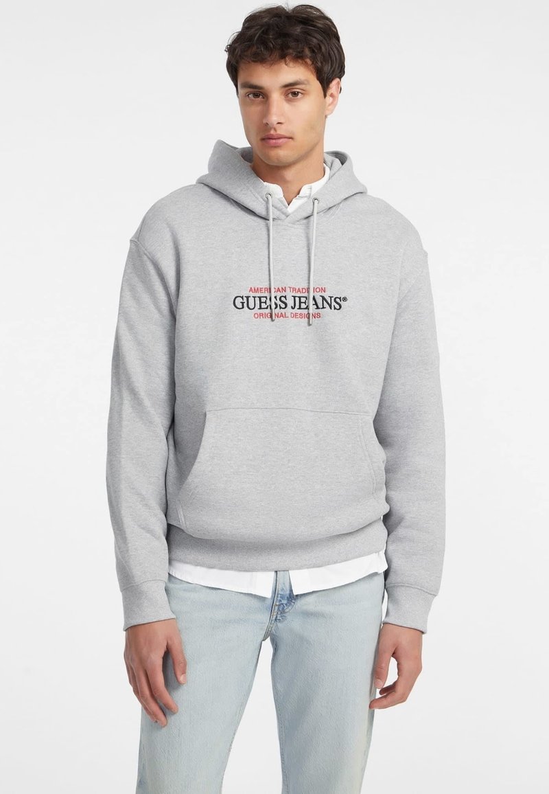 Felpa grigia con cappuccio e coulisse, con un logo nero e rosso sul petto. Indossata sopra una camicia bianca con colletto e abbinata a un paio di jeans azzurri chiari.