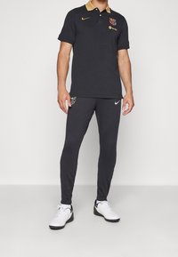 Czarny polo z złotym kołnierzykiem i logo FC Barcelona, w zestawieniu z czarnymi spodniami i białymi sportowymi butami Nike. Prosty design i sportowy krój.