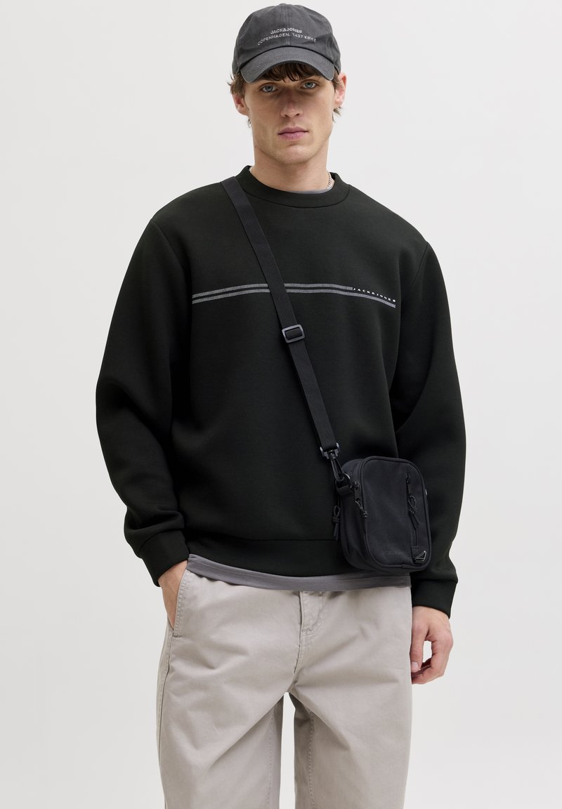 Jeune homme portant un sweat-shirt noir, un pantalon beige, un sac bandoulière noir et une casquette noire avec un texte blanc, debout devant un fond uni.