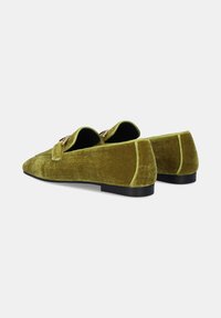 Un par de mocasines de terciopelo verde oliva sin cordones con detalle de hebilla dorada y suelas planas negras sobre un fondo blanco.