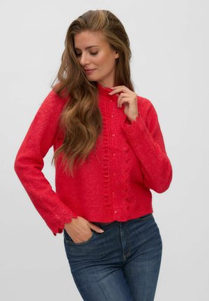 Femme aux longs cheveux ondulés portant un cardigan rouge boutonné et un jean bleu, posant avec une main près du visage et l'autre dans la poche.