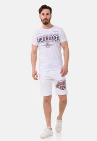 Weißes T-Shirt mit auffälligem grafischen Schriftzug, kombiniert mit weißen Shorts, die mit einem roten und schwarzen Logo-Print versehen sind. Dazu werden weiße Sneakers getragen.