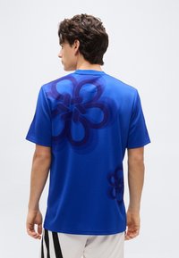 Young man seen from behind wearing a blue short-sleeve shirt with large dark blue floral patterns and white pants with black stripes.