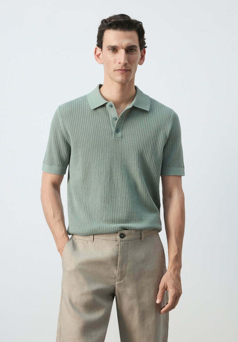 Homme portant un polo en maille vert clair à manches courtes et un pantalon beige, debout avec une main dans la poche sur un fond uni.