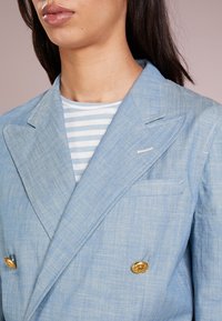 Světle modrý lněný dvouřadý blazer se zlatými knoflíky, špičatými klopami a texturovaným povrchem. Pod ním je vidět pruhované tričko.