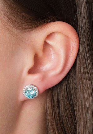 Woman's ear with a round stud earring featuring a large light blue gem surrounded by smaller clear stones.