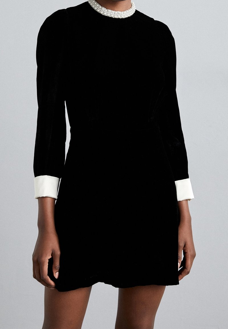 Personne portant une robe noire à manches longues avec des poignets blancs et un collier de perles blanches sur un fond gris uni.