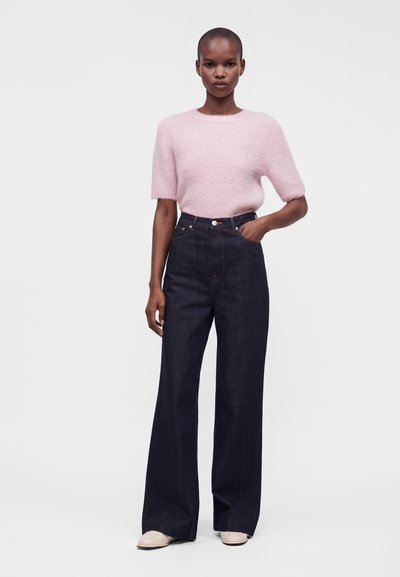 Mujer con un jersey rosa claro de manga corta y textura peluda metido en unos vaqueros oscuros de tiro alto y pierna ancha, con zapatos de color claro.
