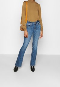 Maglietta marrone a maniche lunghe con scollatura arricciata, abbinata a jeans svasati di media tonalità, e stivaletti neri con punta a punta. Borsa a spalla marrone.