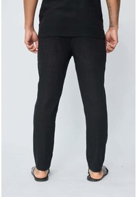 Pantalons noirs en tissu léger, coupe slim avec des jambes droites, portés avec une chemise noire et des sandales, vus de dos.