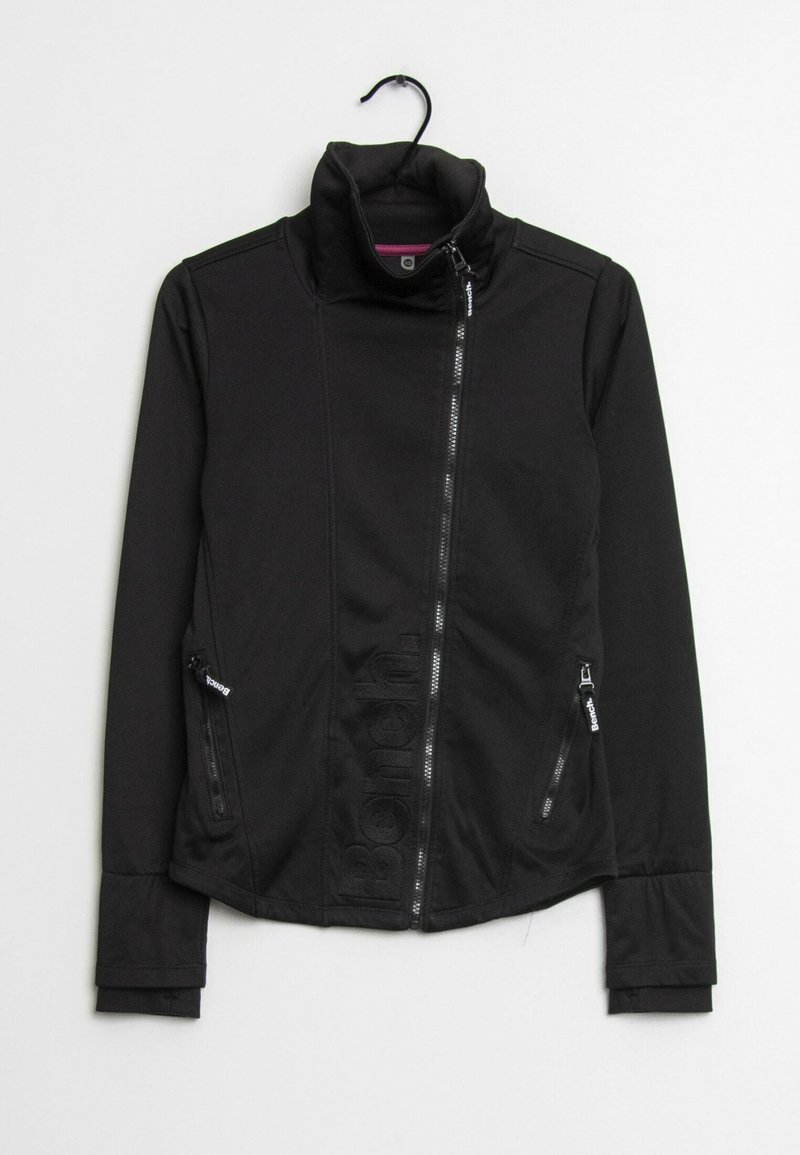 Schwarze Softshell-Jacke mit hohem Kragen. Verfügt über einen Frontreißverschluss, zwei seitliche Reißverschlusstaschen und ein geprägt detailiertes Logo an der Seite.