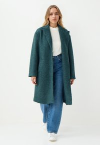 Teal fuzzy jas met een gekarteld kraag, lange mouwen en een open voorkant. Draagt over een witte top en wijde blauwe jeans. Witte sneakers zijn zichtbaar.