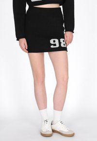 Gonna mini nera lavorata a maglia con design del numero bianco "98". Vita elastica, texture liscia, abbinata a calzini bianchi e sneakers.