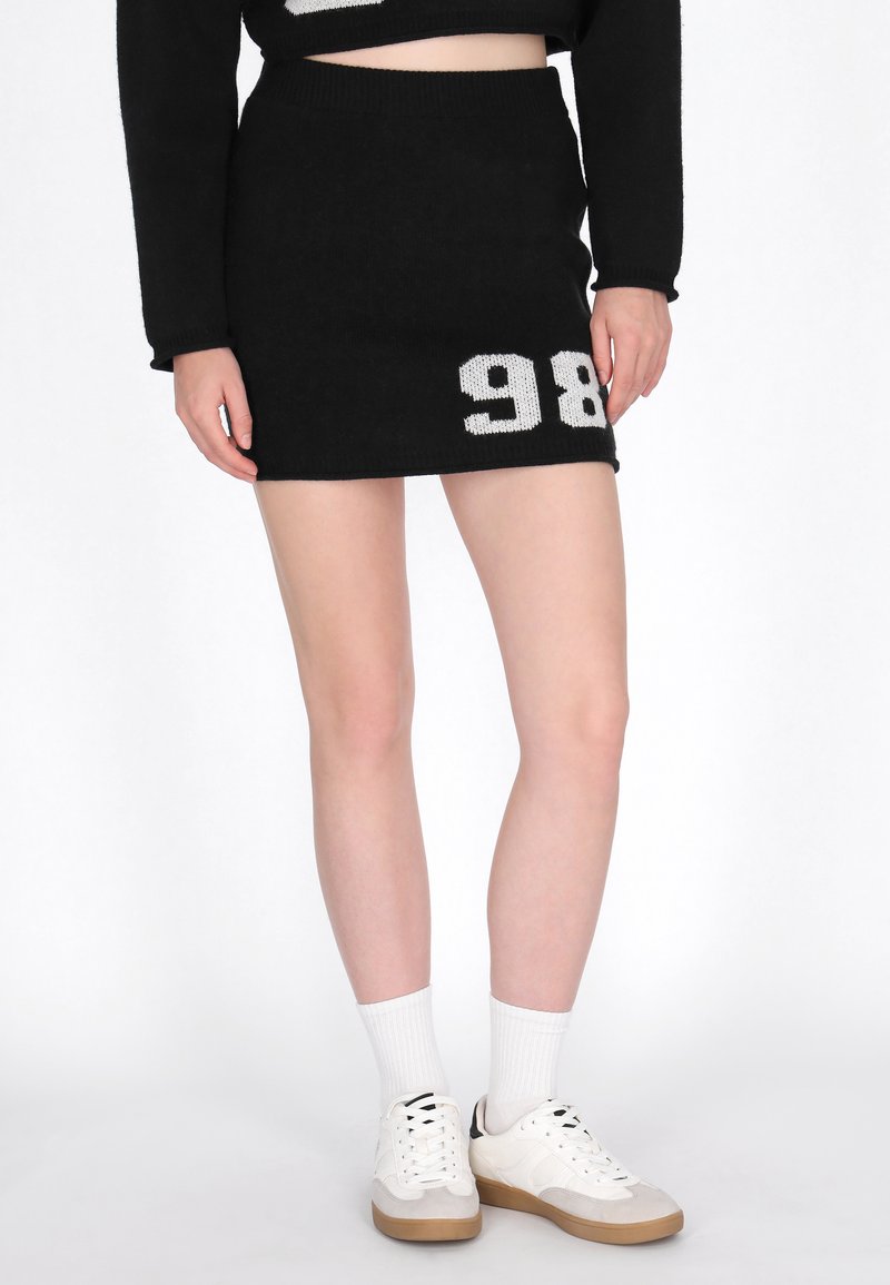 Gonna mini nera lavorata a maglia con design del numero bianco "98". Vita elastica, texture liscia, abbinata a calzini bianchi e sneakers.