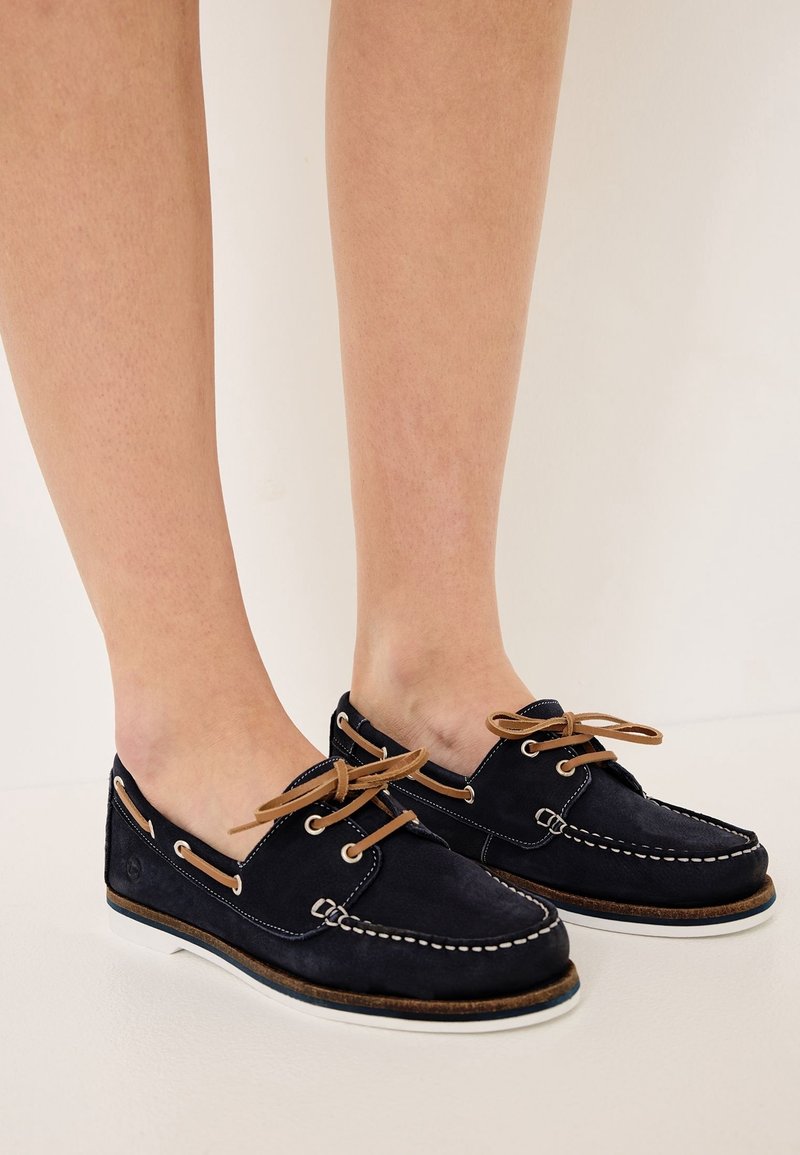 Chaussures bateau en daim bleu marine avec lacets en cuir marron clair et coutures blanches portées pieds nus sur un fond uni.