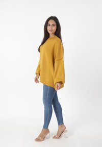 Maglione a maglia oversize color senape con maniche larghe, abbinato a jeans blu aderenti e sandali con tacco trasparenti su uno sfondo semplice.