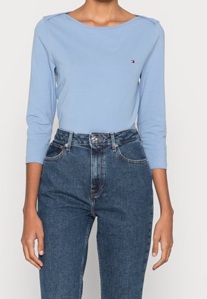 Person trägt ein hellblaues Langarmshirt mit kleinem Logo auf der Brust und hoch taillierte blaue Jeans.