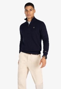 Suéter de cuello con cremallera en azul marino, con una textura de punto suave, combinado con pantalones de color beige claro. Presenta detalles en dorado y puños acanalados en las mangas y el dobladillo.
