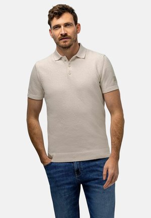 Man met kort bruin haar, die een beige polo met korte mouwen en een blauwe spijkerbroek draagt, staat met één hand in zijn zak.