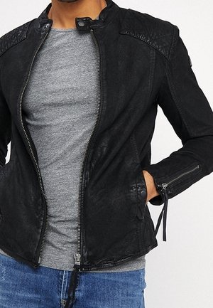 Mężczyzna ubrany w czarną kurtkę na zamek z pikowanymi ramionami na szarej koszuli i niebieskich dżinsach, z jedną ręką w kieszeni kurtki.