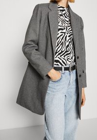 Blazer en espiga gris con solapas muesca, cierre de un solo botón, llevado sobre una blusa de estampado de cebra en blanco y negro y jeans de color azul claro.