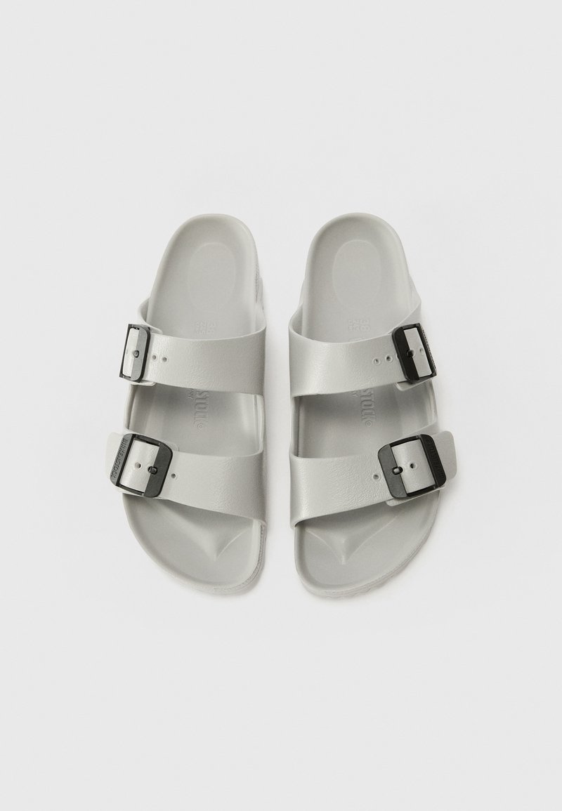 Graue Sandalen mit zwei breiten Riemen, die jeweils eine schwarze Schnalle haben. Glatte, matte Textur und konturierter Fußbett. Flache Sohle.