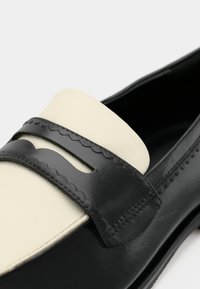 Sapato de couro preto e branco, tipo loafer, com detalhes em forma de onda e uma alça recortada na parte frontal, mostrando um close-up da seção superior frontal.