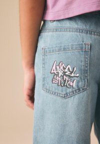 Persoană purtând blugi din denim albastru deschis cu text în stil graffiti pe buzunarul din spate și o cămașă mov, cu mâna odihnindu-se pe lângă corp.