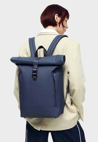 Ritināms mugursoma tumši zilā krāsā. Izgatavota no ūdensizturīga materiāla, aprīkota ar regulējamām siksnām, diviem sānu kabatām ar rāvējslēdzējiem un sprādzi.