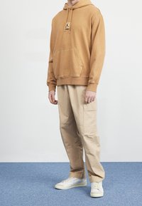Felpa marrone con tasca a marsupio e patch con logo, abbinata a pantaloni cargo beige e sneakers bianche su uno sfondo di tappeto blu.