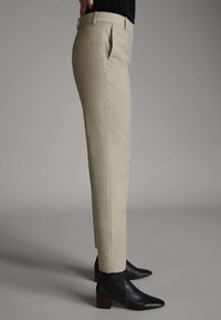 Vue latérale d'une personne portant un pantalon beige sur mesure avec des plis et des bottines noires à talons carrés sur un fond gris.